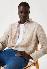 Beige overhemd met lange mouwen en lichte bloemenpatronen, gedragen over een wit T-shirt, gecombineerd met blauwe jeans, zittend op een doorzichtige stoel.