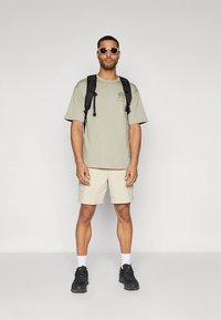 Olivzaļš T-krekls, bēša krāsas šorti, melni sporta apavi, baltas zeķes un melna mugursoma. Aksesuāru komplektā ir saulesbrilles ar biezu rāmi.