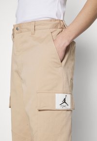Beige cargobyxor med en patch med en svart Jumpman-logotyp, sidofickor och en mjuk tygkvalitet. Justerbar midjedel är synlig.
