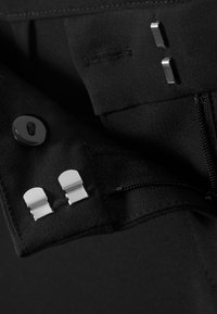 Black fabric with a zipper, button, and metal hardware accents. Two silver clips and stitching detail are visible. Soft texture.