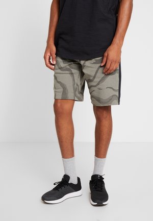 Mann trägt graue Tarnshorts, schwarze Sneakers, graue Socken und ein schwarzes Hemd und steht vor einem einfarbigen Hintergrund.