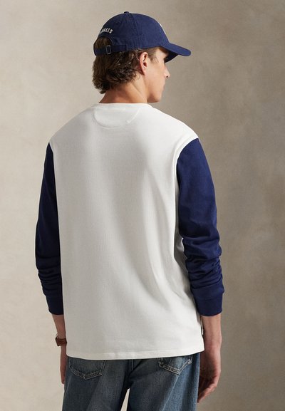 Chemise blanche à manches longues avec des manches bleu marine, texture côtelée et col rond. Portée avec un jean bleu ; la personne est vue de dos.