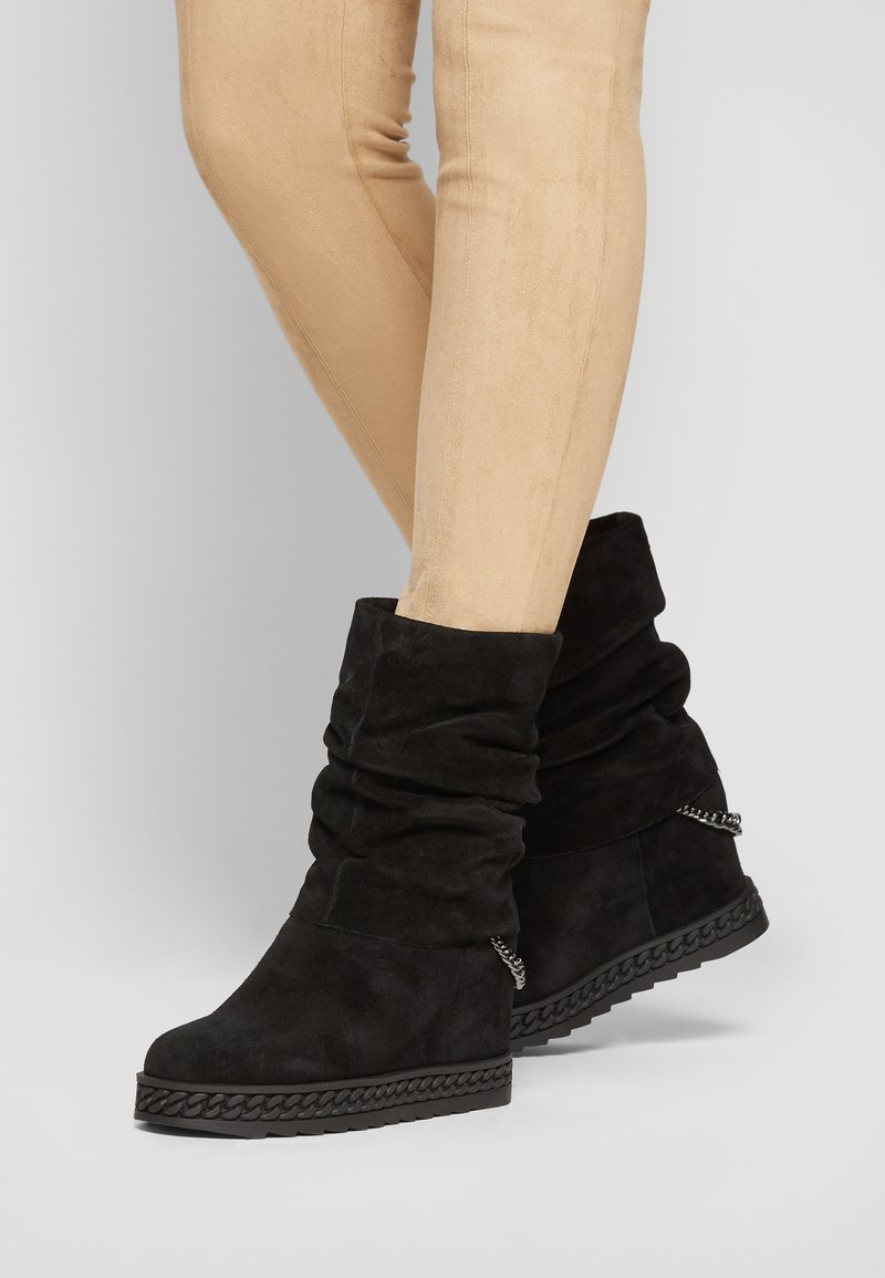 Bottines basses en daim noir décontractées avec détail de chaîne sur la semelle, portées avec un pantalon beige ajusté sur un fond uni.