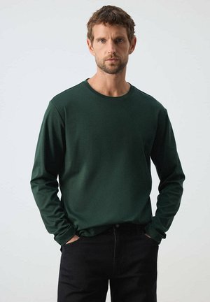 Hombre con pelo corto y castaño que lleva una camisa de manga larga verde oscuro y pantalones negros, de pie con las manos en los bolsillos frente a un fondo liso.