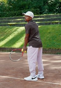 Tenisový hráč drží raketu a tenisové loptičky, má na sebe hnedú polokošeľu, svetloružové nohavice a biele tenisky Nike, na antukovom kurte.