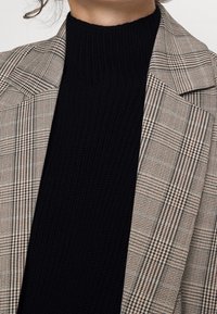 Blazer mit beige-braunem Karomuster, ausgestattet mit einem strukturierten Design. Darunter ein schwarzer Strickpullover mit hohem Kragen und texturiertem Finish.