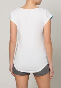Camiseta blanca de manga corta con dobladillo curvado, que presenta detalles en turquesa a los lados. Fabricada en un tejido suave y ligero, con una textura lisa.