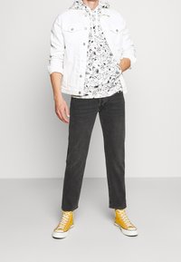 Giacca di denim bianca sopra una felpa con cappuccio a motivi bianco e nero, abbinata a jeans neri e sneaker alte gialle.