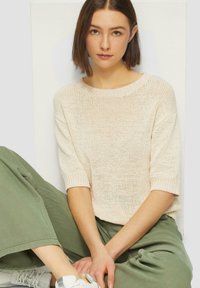 Vaaleanbeige neule, jossa on lyhyet hihat ja leveä pääntie, yhdistettynä oliivinvihreisiin leveälahkeisiin housuihin. Sujuva kankaan pinta.