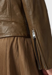 Giubbotto di pelle marrone con finitura liscia, dotato di polsini con zip e design slanciato. Il tessuto è morbido con una leggera lucentezza.