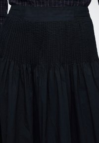 Jupe noire avec des plis froncés et une large ceinture smockée portée avec une chemise à carreaux sombre visible en haut.