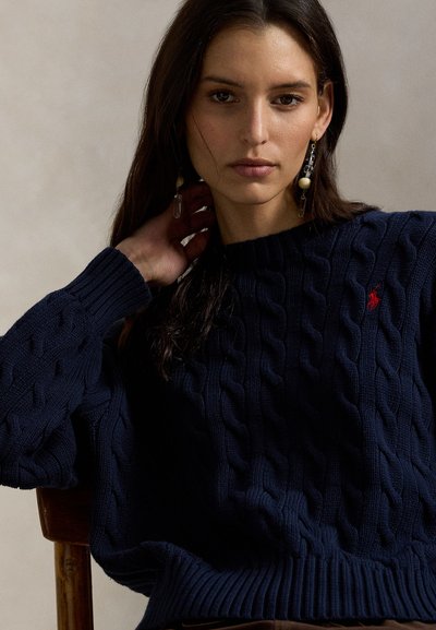 Πλεκτό πουλόβερ σε ναυτικό μπλε με ριμπ μανσέτες και τελείωμα. Διαθέτει μικρό κόκκινο κεντητό λογότυπο. Φοριέται με κρεμαστά σκουλαρίκια.