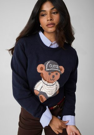 Donna che indossa un maglione blu navy con un orsetto in maglione bianco e cappellino nero con etichetta "GUESS," stratificato sopra una camicia a quadri blu.