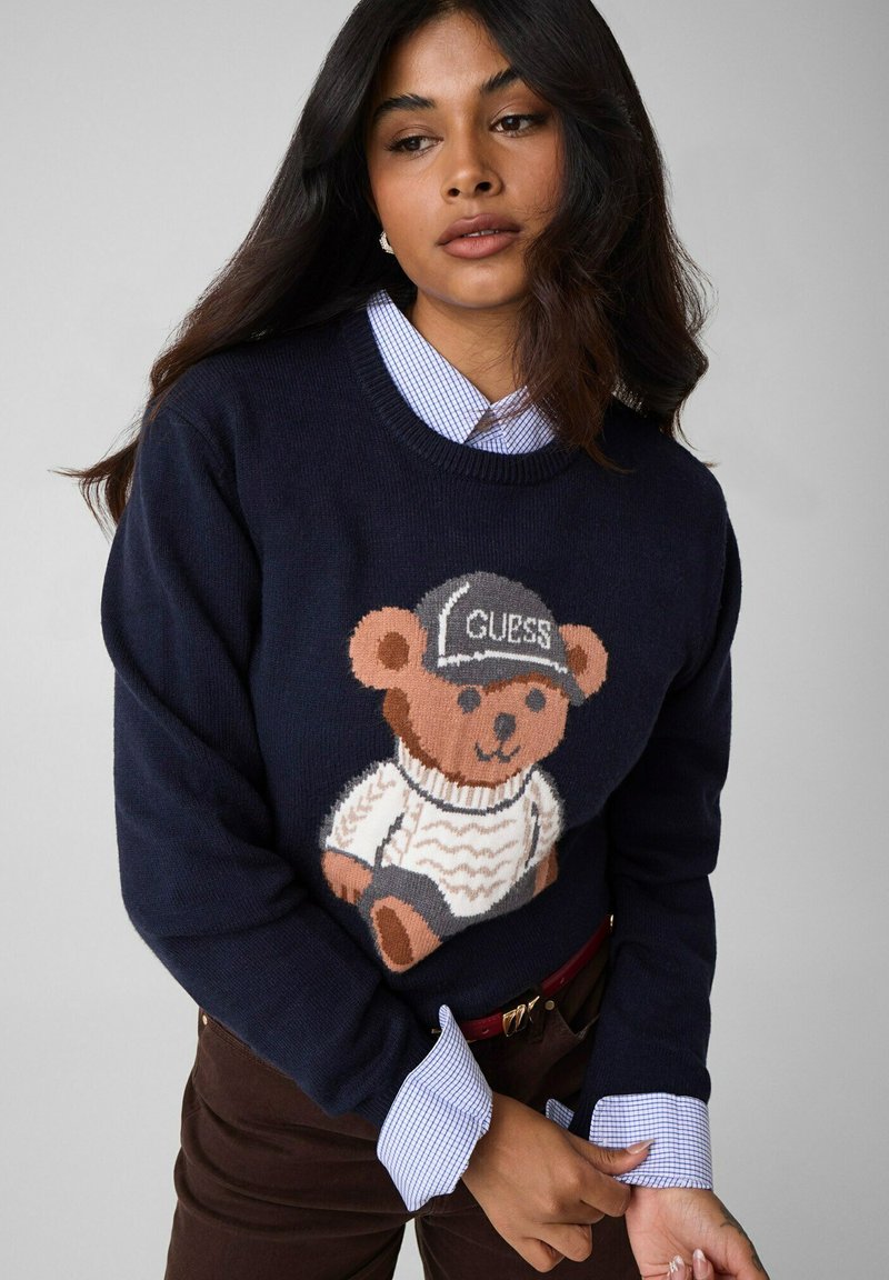Femme portant un pull bleu marine avec un ours en peluche en pull blanc et une casquette noire portant l’étiquette "GUESS," superposé à une chemise à carreaux bleue.