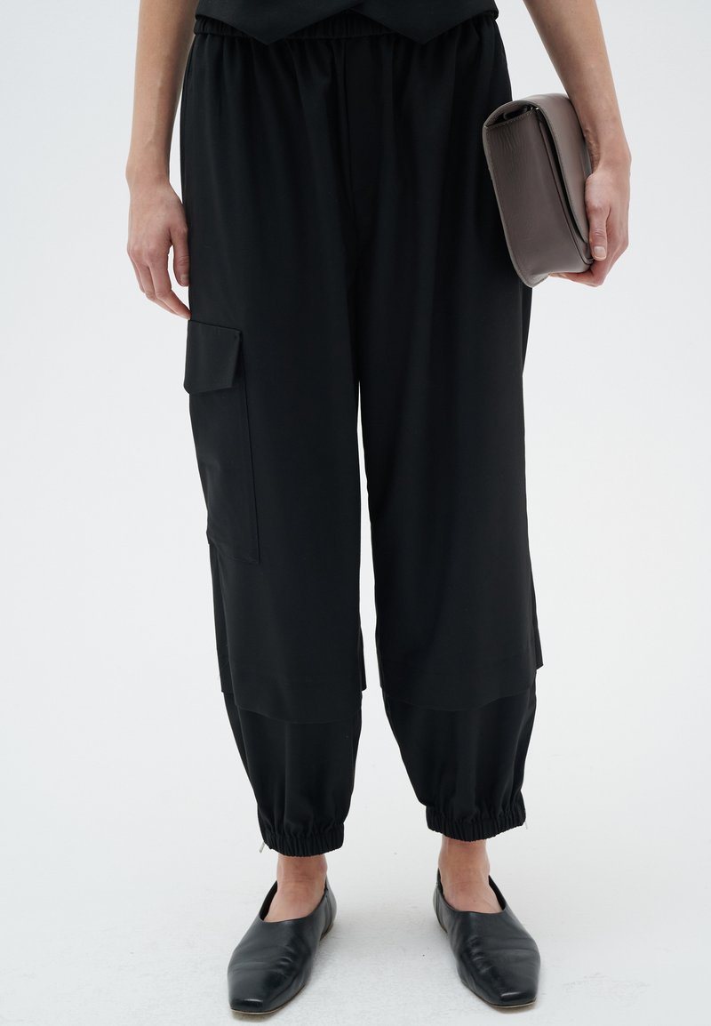 Schwarze Cargo-Hosen mit einem elastischen Bund, Bündchen an den Säumen und Seitentaschen. Kombiniert mit einer schlichten grauen Clutch und schwarzen Flats.