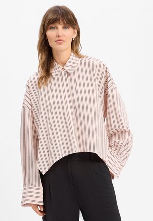 Femme portant une chemise ample à manches longues rose clair avec des rayures noires verticales et un pantalon noir taille haute sur fond blanc.