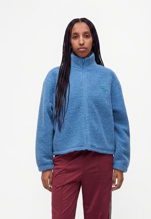 Kvinde med lange fletninger iført en blå fleece lynlåsjakke og bordeaux bukser, stående mod en ensfarvet hvid baggrund.