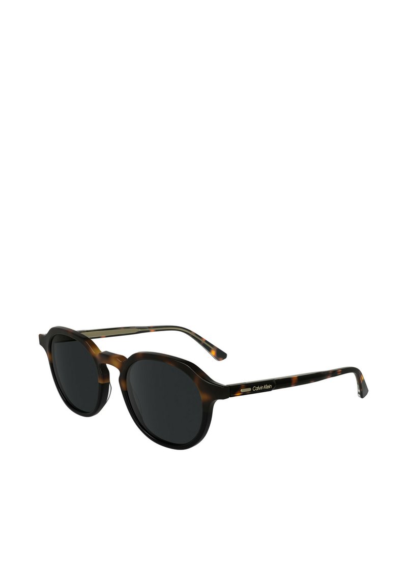 Occhiali da sole rotondi con montatura in tartaruga, lenti scure e dettagli in metallo nero. Le astine presentano il logo argentato di Calvin Klein.