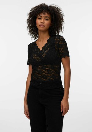 Femme aux cheveux bouclés portant un chemisier noir en dentelle à manches courtes et un pantalon noir, debout devant un fond blanc uni.