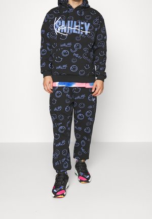 Personne portant un sweat à capuche noir et un pantalon avec un motif de visage souriant bleu et des textes, associé à des baskets noires colorées.