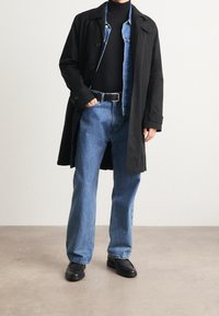 Abrigo negro largo sobre un jersey de cuello alto negro, chaqueta de mezclilla azul clara, vaqueros azules holgados y zapatos negros, de pie sobre una superficie lisa.