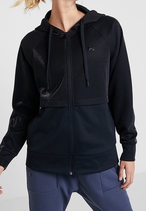 Kobieta ubrana w czarną bluzę na zamek z kapturem i sznurkami oraz ciemnoniebieskie spodnie, stojąca na tle jednolitego szarego tła.