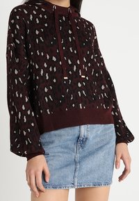Bordeaux trui met zwart-wit luipaardprint, voorzien van een geribde kraag en verstelbare trekkoorden, gedragen over een denim mini rok.