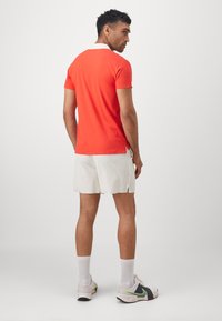 Polo rosso con colletto bianco, abbinato a pantaloni corti bianchi. Il modello è in piedi con una leggera svolta, mostrando il tessuto testurizzato e i dettagli dell'orlo.