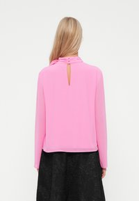 Blusa rosa a maniche lunghe con scollo arricciato e dettaglio dei bottoni sul retro, caratterizzata da un piccolo taglio e una texture di tessuto liscia.