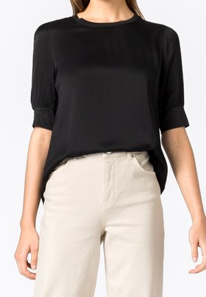 Vrouw die een zwarte blouse met korte mouwen draagt, ingestopt in een beige broek met hoge taille, staand met ontspannen armen langs het lichaam.