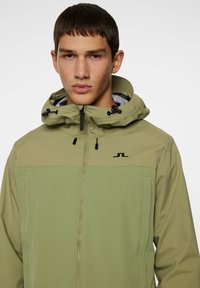 Grøn zip-up jakke med hætte, der har et to-tone design, en glat tekstur og et sort logo på brystet. Justerbare snore i hætten.
