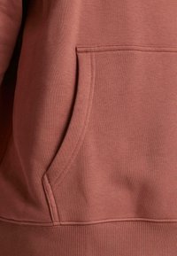 Rustfarvet sweatshirt med frontlomme, glat tekstur og ribbet kant. Syninger er synlige langs lommen og sømmene.