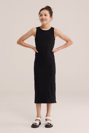 Fată tânără cu aparat dentar, purtând o rochie midi neagră fără mâneci și sandale albe, stând cu mâinile pe șolduri pe un fundal simplu.