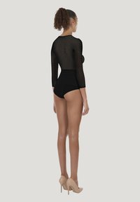 Sort bodystocking i netstof med trekvartlange ærmer, tætsiddende design, gennemsigtig tekstur og en ensfarvet sort bund. Bæres med beige højhælede sko.