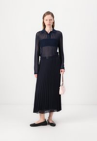 Fustă plisată bleumarin și bluză transparentă cu mâneci lungi și guler. Modelul ține o mică poșetă roz deschis. Pantofi negri cu talpă joasă. Design minimalist.