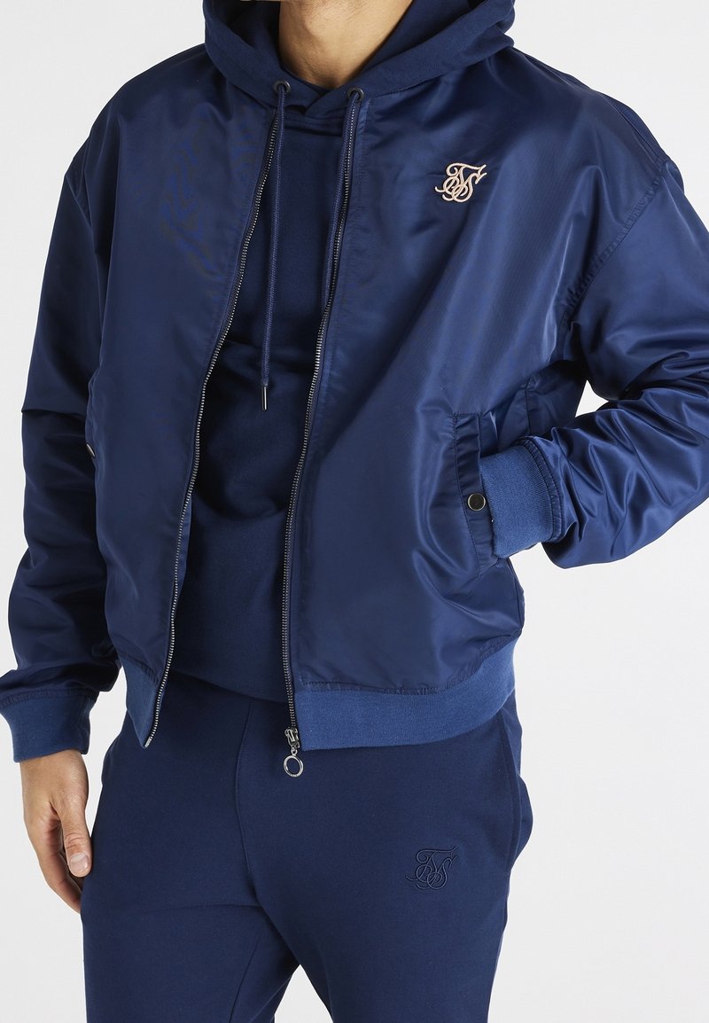 Mörkblå bomberjacka i glänsande tyg med dragkedja fram, ribbade muddar och en diskret logotypsbroderi på bröstet.