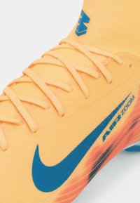 Close-up van een oranje Nike sportschoen met een blauwe swoosh-logo, "AIR ZOOM" tekst en oranje veters op een lichte achtergrond.