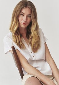 Hvid linned kortærmet skjorte med krave og tre brune knapper, parret med lyse shorts. Baggrunden er neutral.