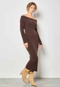 Bruine geribbelde gebreide off-shoulder jurk met lange mouwen, aansluitende silhouet en uitlopende zoom, gecombineerd met beige losse enkellaarzen.