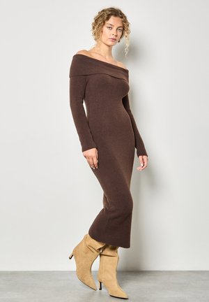 Bruine geribbelde gebreide off-shoulder jurk met lange mouwen, aansluitende silhouet en uitlopende zoom, gecombineerd met beige losse enkellaarzen.