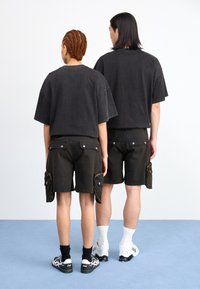 T-shirts oversized pretos combinados com calções cargo escuros com bolsos laterais. Ambos usam ténis atléticos brancos e pretos. Fundo simples.