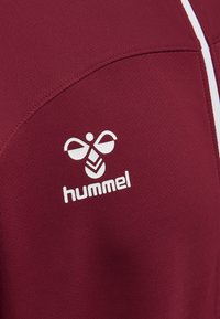 Bordeaux sportsjersey med en struktureret overflade, med et fremtrædende hvidt Hummel-logo på den øverste venstre side. Hvide detaljer langs sømmene.