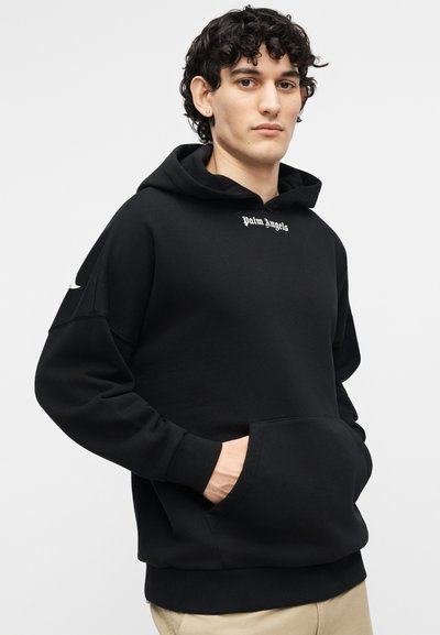 Noor mees lokkis juustega, kes kannab musta Palm Angels kapuutsi ja paneb käed esitaskusse, seistes ühevärvilise hele tausta ees.