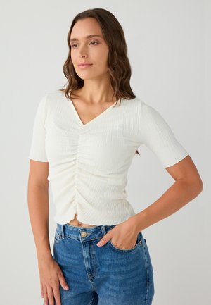 Donna con capelli castano ondulati indossa una maglietta bianca a maniche corte con arricciature e jeans blu, in piedi con una mano in tasca su uno sfondo neutro.