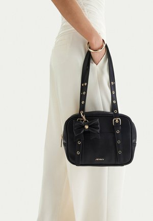 Mujer con conjunto blanco sosteniendo un bolso negro rectangular con detalle de lazo y herrajes dorados en la correa y la cremallera.