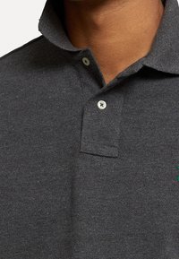 Nahaufnahme einer Person, die ein dunkelgraues Poloshirt mit umgeschlagenem Kragen und zwei sichtbaren weißen Knöpfen trägt.