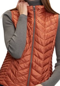 Zbliżenie na osobę noszącą kamizelkę z pikowanego materiału w kolorze rdzawym z suwakiem z przodu, na szarym, prążkowanym, długim rękawie.