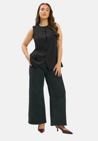 Mujer con el cabello largo, vistiendo una blusa negra sin mangas con botones, pantalones anchos negros y tacones de punta burdeos, de pie con una mano en el bolsillo.