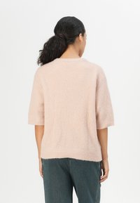 Light pink, short-sleeved sweater with a textured, ribbed design. Soft material, rounded neckline, and relaxed fit. Worn with dark blue jeans.