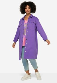 Paarse button-up jas met lange mouwen, twee borstzakken, gecombineerd met een roze graphic t-shirt en lichtblauwe cropped jeans. Witte sneakers.